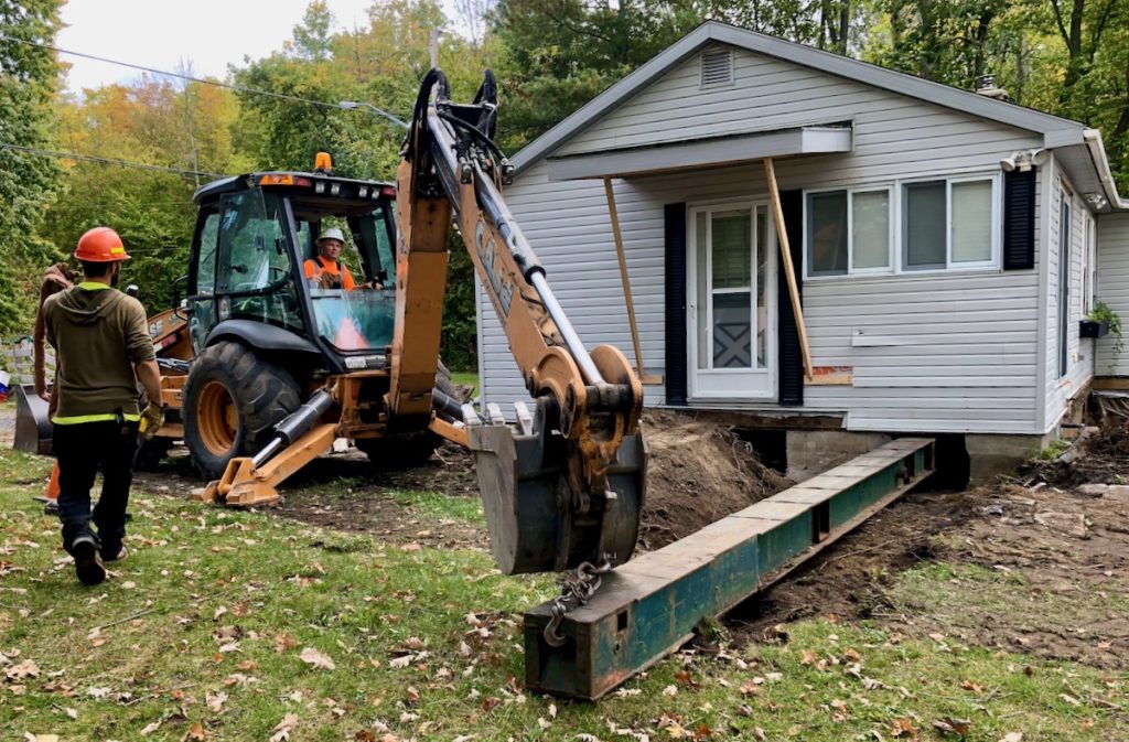 Installing steel beams with backhoe under house through hole in foundation